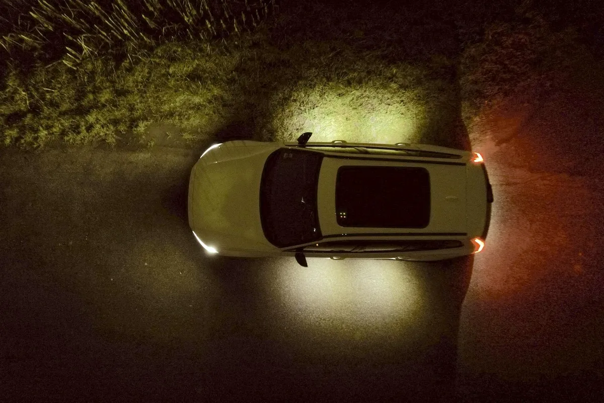 Overhead nighttime view of a white car parked on a dark road, illuminated by its headlights and taillights, with green vegetation visible along the left side.