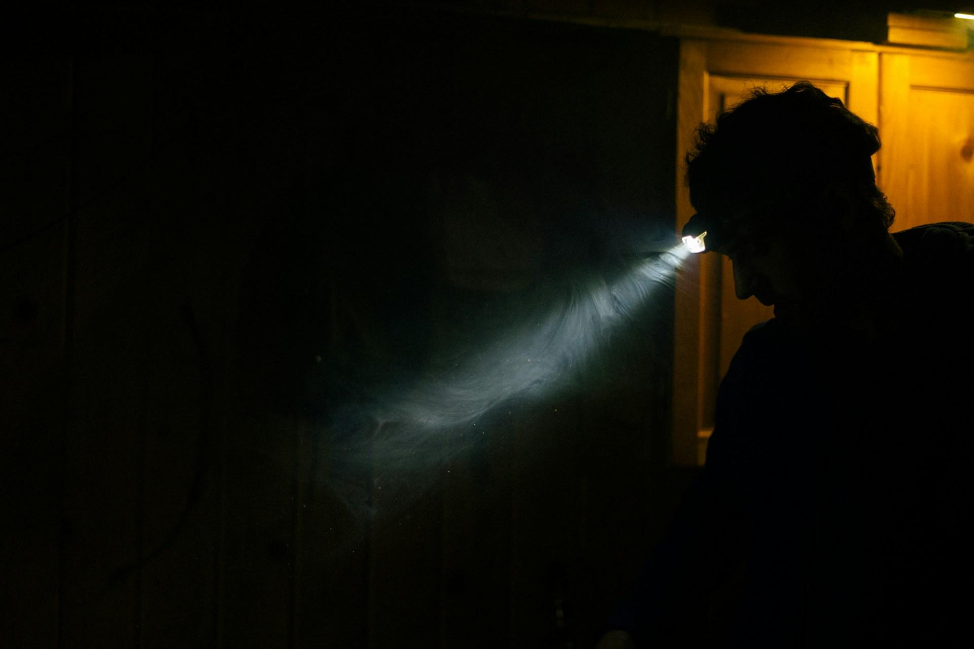 A man in profile investigates a dark attic, his headlamp cutting a single beam of light through the gloom and revealing motes of dust in the air.