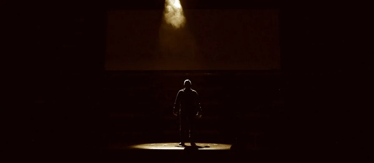 A lone figure illuminated by a single spotlight on an empty stage, surrounded by darkness.
