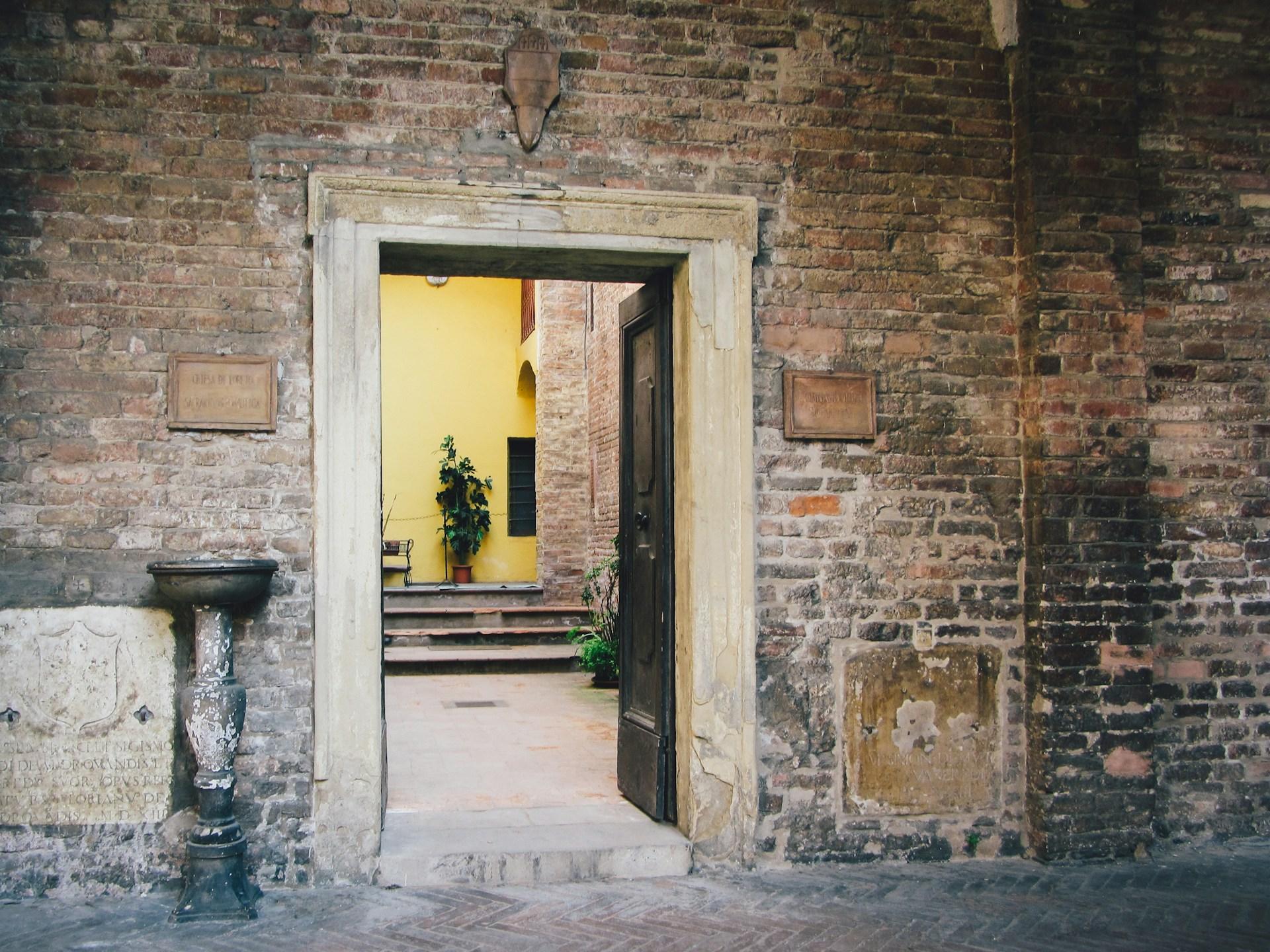 A slightly open wooden door in a rustic brick wall, with warm yellow light streaming through from the room inside.