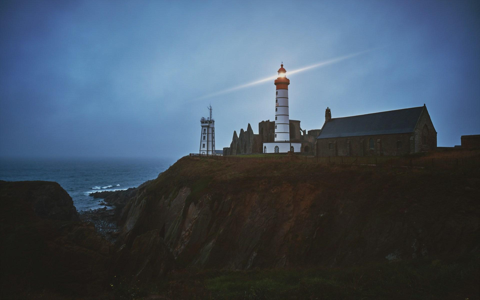 A sturdy lighthouse stands firm on a dark, rocky coast, its powerful beam cutting through the fog at dusk.