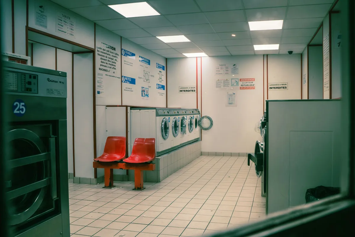 An empty laundromat with tiled floors and fluorescent lighting. Red plastic chairs sit in the middle of the room. The overall atmosphere is clean but slightly worn and quiet.