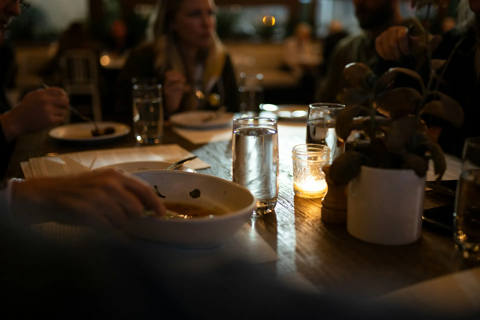 A candlelit dinner table at night, with a hand reaching for a bowl and blurred figures in the background.